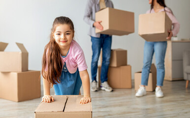 House moving concept. Young family of three relocating and unpacking boxes in new flat indoors. Little cute girl pushing cardboard box and smiling to camera, parents talking on background