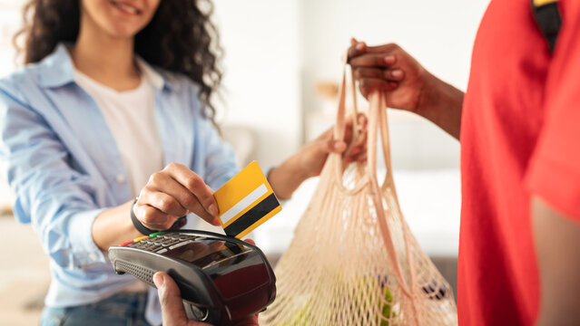 Digital Financial Transaction. Closeup of woman using credit card for contactless payment, black deliveryman holding Point Of Sale terminal in hand, giving string bag with groceries, selective focus
