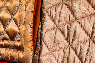 Close-up of ornate quilted fabrics in rich tones and baroque floral patterns hanging in sunlight. Maximalist decor, secondhand textiles, brocade revival, retro fabric trends