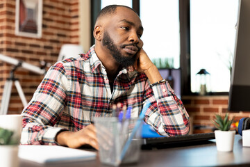 African american businessman sits at desk in home office, leaning on table with tired expression,...