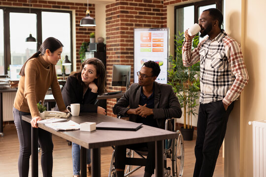 Male and female business partners engage in a casual yet meaningful discussion at office desk, promoting teamwork. Group of coworkers chatting with each other while some enjoy a cup of coffee.