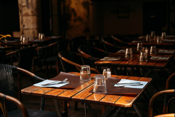 Wooden tables with glasses and cutlery set for customers on a shaded cafe terrace. Terrace dining, slow coffee culture, local eateries, mindful lifestyle