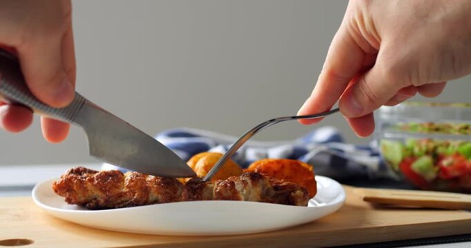Hands using knife and fork cutting a piece of grilled chicken breast served with roasted potatoes on a white plate and wooden board, with a fresh salad on background