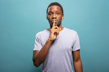 Black man standing against a blue background, pressing index finger to lips in a hush gesture,...