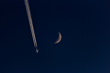 A plane in the night sky with the moon and contrail.