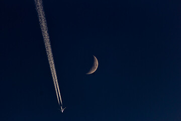 A plane in the night sky with the moon and contrail.