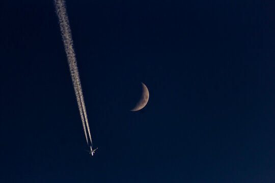 A plane in the night sky with the moon and contrail.