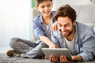 Free time together. Father and son playing online on digital tablet, boy pointing finger to screen,...