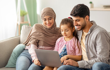 Young muslim parents and girl using laptop together at home, surfing internet or having video chat...