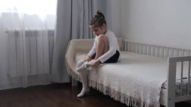 Side view of a young girl sitting on a bed and putting on tights in a bedroom at home.