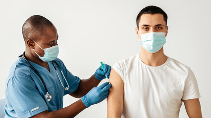 Antivirus program for population against covid-19 outbreak, vaccination campaign. Adult african american doctor make injecting young patient in arm isolation on gray background, empty space