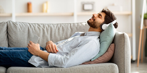 Obraz premium Happy arab guy in wireless headphones lying on comfy couch with closed eyes and listening to music at home, copy space, side view. Man spending lazy time on weekend, enjoying his favorite song