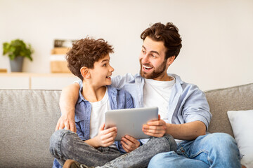 Family weekend. Excoted dad and son using tablet computer, surfing internet while spending time...