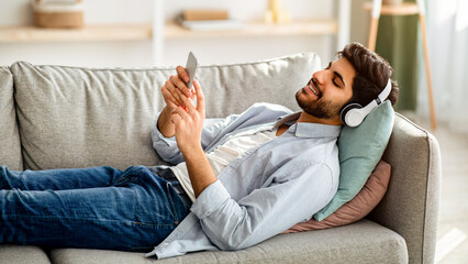 Free time. Relaxed arab guy listening to music in wireless headphones, lying on comfy couch on weekend at home. Man chatting on mobile phone and enjoying favorite songs