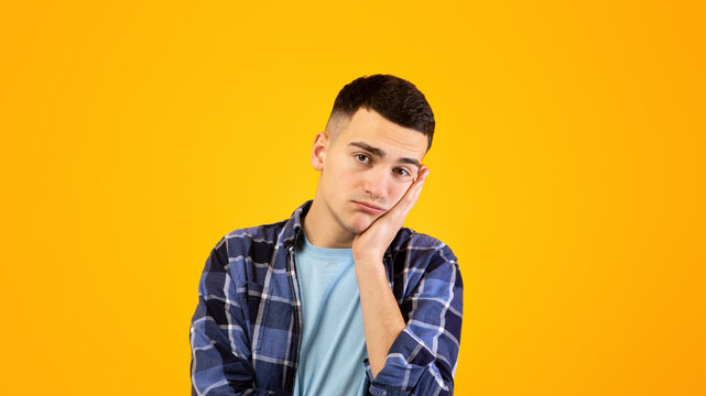 Portrait of young guy feeling bored, leaning on his hand, having dull day on orange studio background. Funky Caucasian man being annoyed or disappointed, showing lack of interest in something
