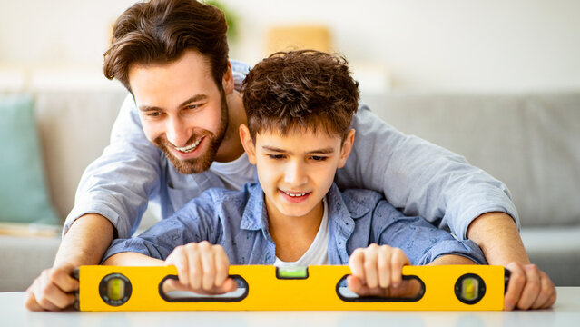 Happy father teach son using level tool for measure surface, doing apartment repair together themselves. Family doing hobby together, home makeover or renovation concept