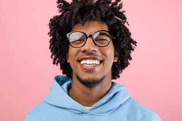 Portrait of joyful black teenager with pleasant smile wearing glasses, looking at camera on pink...