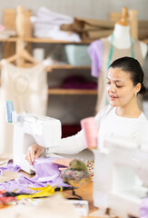 Attentive woman tailor sewing textile using sewing machine sitting at workplace in small fashionable sewing atelier