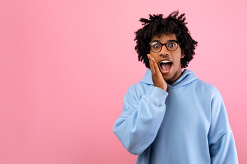 Cool African American teen in glasses opening mouth in shock, shouting WOW, touching cheek, looking...