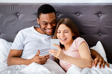 Online call, mobile app, remote shopping concept. Happy young Caucasian female and her black boyfriend looking at smartphone screen, chatting in social networks, searching internet
