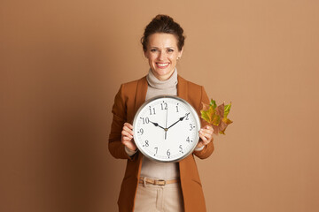 A smiling woman in business casual attire holds a wall clock and autumn leaves, symbolizing time...