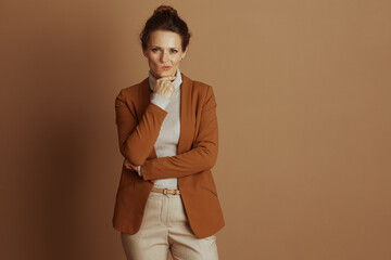 Fototapeta premium A thoughtful businesswoman, hand on chin, gazes at camera, pensive against a plain brown background. Her beige turtleneck and open rust-colored blazer suggest contemplation or questioning.