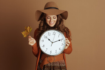 A smiling woman with curly brown hair and a wide-brimmed hat holds a clock and a fall leaf,...