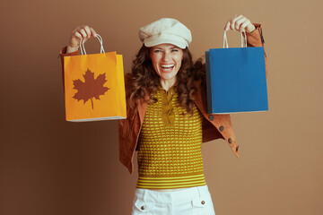 A joyful woman in a beige cap and brown suede jacket proudly holds up autumn shopping bags, one...