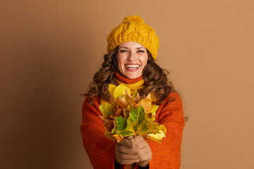 A smiling woman, wearing yellow knit hat and orange sweater, holds bouquet of vibrant autumn...