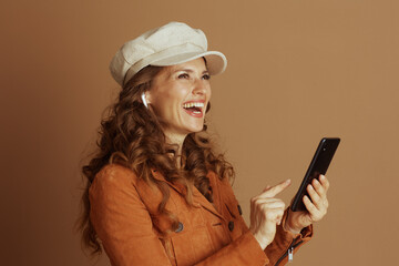 A smiling woman in an orange suede jacket and newsboy cap gestures towards her smartphone screen,...