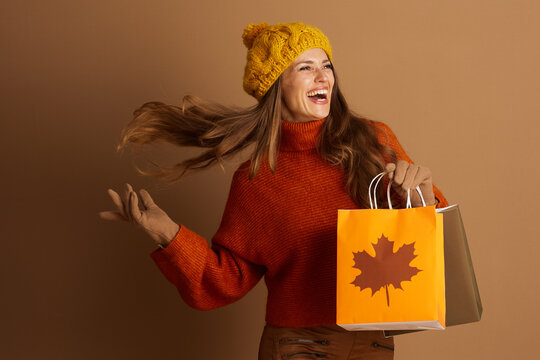 A joyful woman in a yellow pompom hat and orange turtleneck laughs, her hair playfully blowing as she holds autumn-themed shopping bags. The scene captures the excitement of seasonal consumerism.