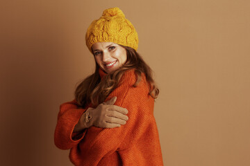 A smiling woman with long brown hair, wearing a yellow pompom beanie and an oversized orange...