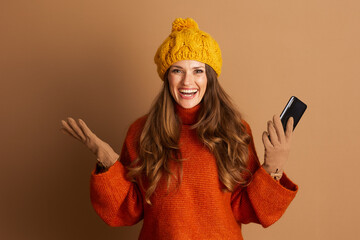 A cheerful woman in pom-pom hat and orange turtleneck sweater smiles brightly, holding smartphone...