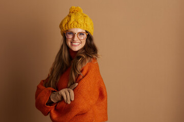 A smiling woman in an orange turtleneck, yellow beanie, and glasses stands with arms crossed,...