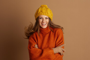 Cheerful woman in orange turtleneck and yellow cable knit beanie smiles broadly, her hair slightly...