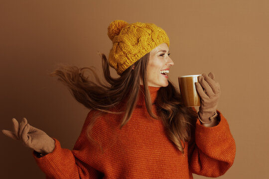 A cheerful woman in yellow beanie and orange turtleneck smiles broadly, holding ceramic mug. Her long hair flows. The warm brown background enhances the cozy, joyful atmosphere for autumn or winter.