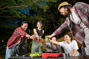 Indonesian southeast asian people relaxing together outdoors and enjoying refreshing drinks while cooking sliced beef on a grill pan. The scene of the lifestyle of enjoying food in a natural setting