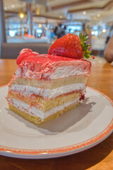 Close-up of a layered strawberry cream cake with a fresh strawberry on top, set on a white plate against a warm, cozy cafe background.