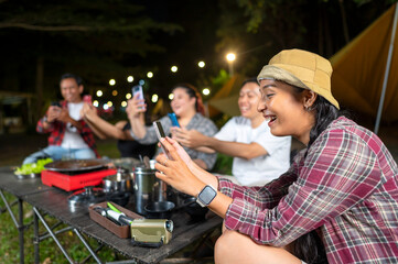 Indonesian southeast asian people enjoying a camping at night, using their cellphones to capture moments and share experiences. The scene of the lifestyle of enjoying camping in a natural setting