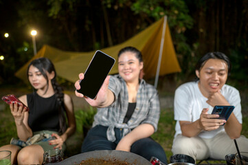Indonesian southeast asian people enjoying a camping at night, using their cellphones to capture moments and share experiences. One of them showed her cellphone display. Mockup or Copy space