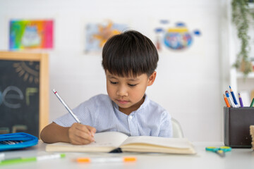 Asian boy studying with pencil at home smiling writing with notebook and school supplies, online...
