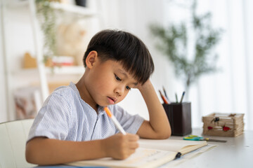 Asian boy studying with pencil at home smiling writing with notebook and school supplies, online...