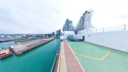A ground-level fisheye view of a large cruise ship docked at a container terminal under a cloudy...