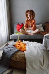 Young woman with curly hair sitting on a sofa, surrounded by colorful clothes, showcasing a casual...