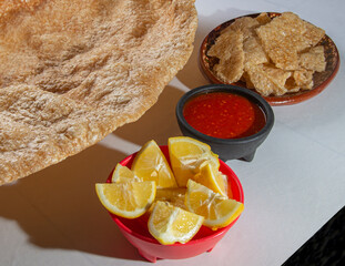 Pork skin or chicharron with lemon slices and spicy red salsa on the side on a white table 45 angle...