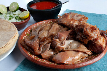 Mexican cuisine, a dish called CARNITAS on a clay plate with pork rinds and belly with onions and very spicy red sauce with corn tortillas on one side.