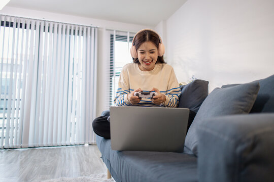 A young woman sits on a sofa wearing headphones while holding a game controller and playing a video game on her laptop. Her cheerful expression shows enjoyment and excitement.