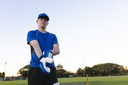 Male cricketer pulling on wicket-keeping gloves by stumps on cricket field in blue jersey, cap
