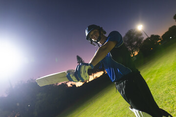 Male cricketer standing on field under floodlights in batting gear holding bat, copy space