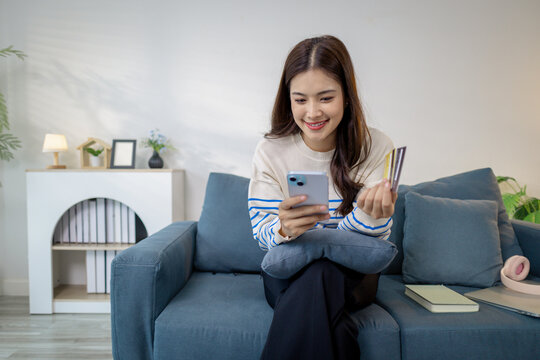 A young woman sitting on a sofa at home smiling while holding a smartphone in one hand and a credit card in the other, representing online payment, digital finance, and e-commerce lifestyle.